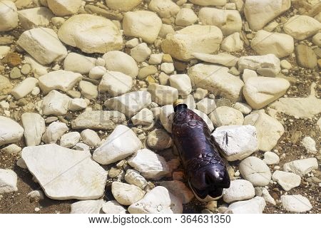 Dark Plastic Bottle On The Rocky Shore Of The River, Sea. Concept Of Pollution Of Rivers, Seas And O