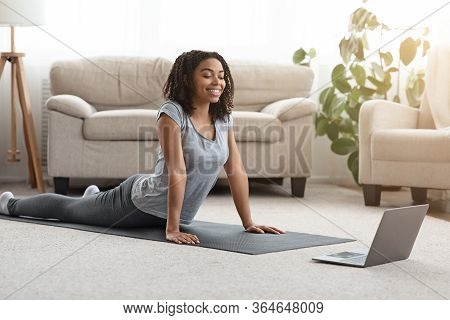 Young Black Woman Practicing Yoga At Home With Laptop, Watching Online Tutorials, Excersising In Liv