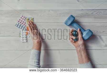 Senior Wrinkled Hand Pushing Back Pills And Thermometer, Dumb-bell On A Table. Another Old Woman Han