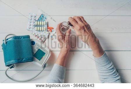 Senior Hands With Medicine Bottle Putting Drops In A Glass, Pills, Tonometer, Thermometer On A Table