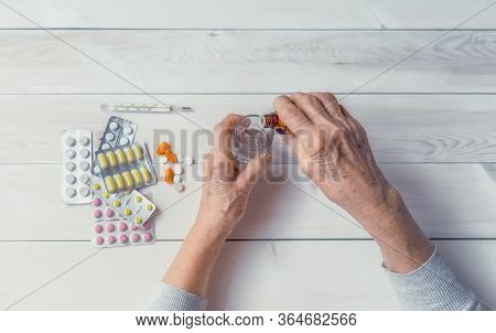 Senior Hands With Medicine Bottle Putting Drops In A Glass, Pills, Tonometer, Thermometer On A Table