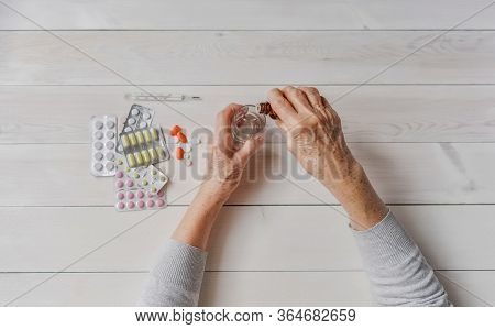 Senior Hands With Medicine Bottle Putting Drops In A Glass, Pills, Tonometer, Thermometer On A Table
