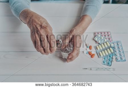 Senior Hands With Medicine Bottle Putting Drops In A Glass, Pills, Tonometer, Thermometer On A Table