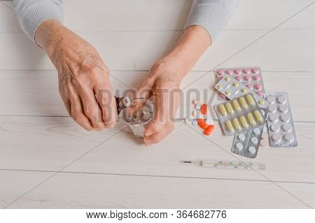 Senior Hands With Medicine Bottle Putting Drops In A Glass, Pills, Tonometer, Thermometer On A Table