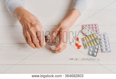 Senior Hands With Medicine Bottle Putting Drops In A Glass, Pills, Tonometer, Thermometer On A Table