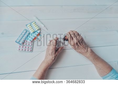 Senior Hands With Medicine Bottle Putting Drops In A Glass, Pills, Tonometer, Thermometer On A Table