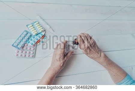 Senior Hands With Medicine Bottle Putting Drops In A Glass, Pills, Tonometer, Thermometer On A Table