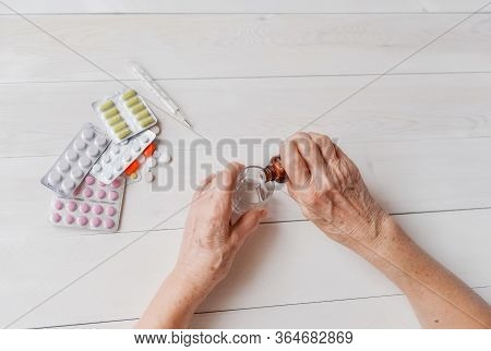 Senior Hands With Medicine Bottle Putting Drops In A Glass, Pills, Tonometer, Thermometer On A Table