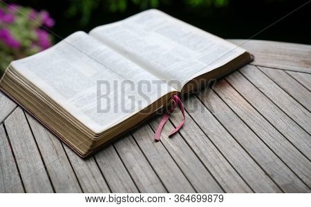 Open Bible On Wooden Table. Soft Blur Effect With Focus On Book Mark. Garden View At Background.