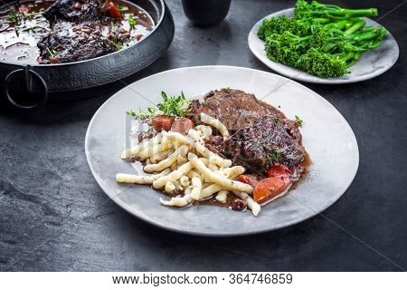 Traditional German braised beef cheeks in brown red wine sauce with noodles and broccoli offered as closeup on a modern design plate and stewpot on an old rustic board 