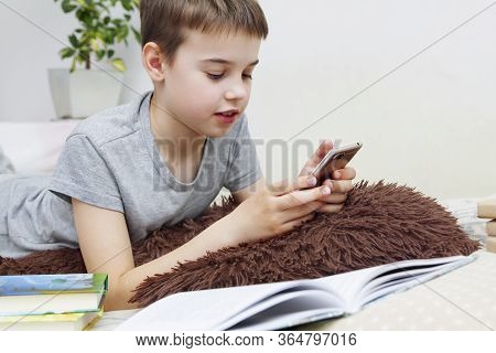 A Boy Is Lying On The Bed With A Book And Smartphone. Home Schooling. Quarantine Coronavirus Isolati