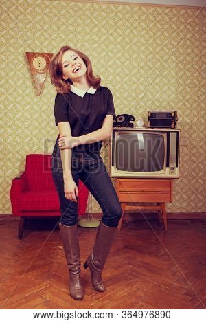 Young smiling ecstatic woman looking at camera and happy smiling in room with vintage wallpaper and interior, retro stylization 60-70s. Furniture, tv set and another technique of the mid 20th century