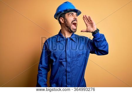 Mechanic man with beard wearing blue uniform and safety helmet over yellow background shouting and screaming loud to side with hand on mouth. Communication concept.