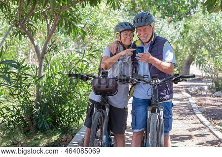 Active Caucasian Senior Couple With Electrobikes Standing Outdoors In The Park Looking At Cellphone.