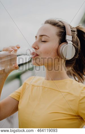 Young Woman Drinking Water And Listening Music Trough Headphones, During Jogging In City, Healthy Li