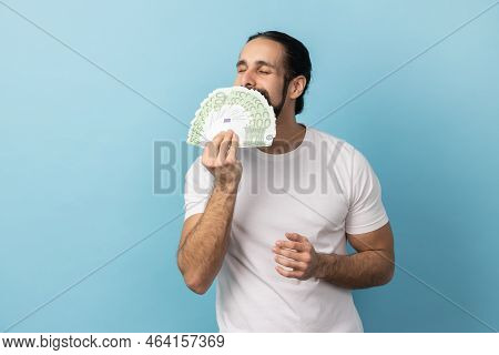 Portrait Of Avaricious Rich Man With Beard Wearing White T-shirt Smelling Earned Euro Banknotes, Enj