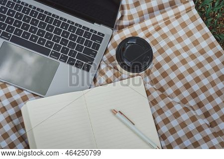 Work On Laptop On Picnic Blanket On The Lawn In The Park- Next To A Headphone, Notebook  And Cup Of 