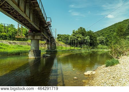 The Old Railroad Bridge Over The River. The Clear Water Of The Shallow River. Blue Sky. Green Forest