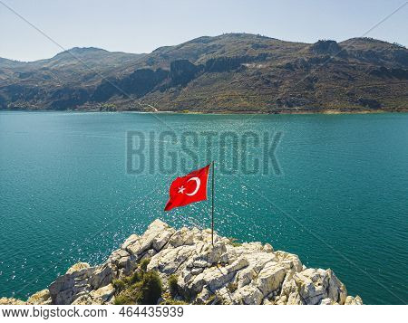 Vivid Red Turkish Flag On Rocky Shore Zone Of Turquoise Oymapinar Lake In Turkey. Summertime Destina