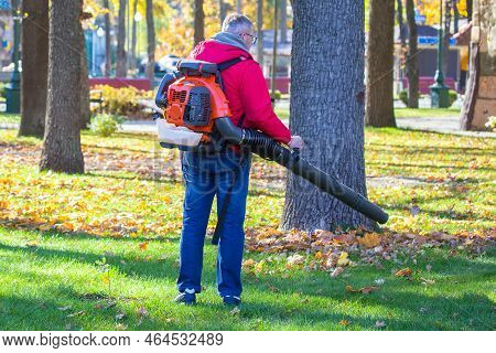 Working In The Park Removes Leaves With A Blower. Park Cleaning Service. Removing Fallen Leaves In A