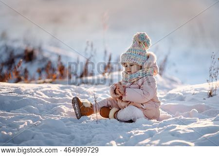 An Adorable Little Girl In A Warm Coat And A Colorful Hat With A Pom-pom Stands In The Snow On The S