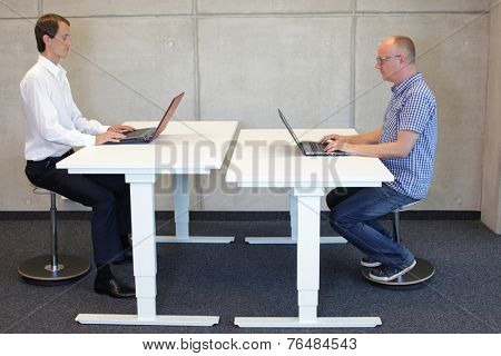 two men  coworking in correct sitting posture on pneumatic leaning seats  with laptops  at electric height adjustable desks in office