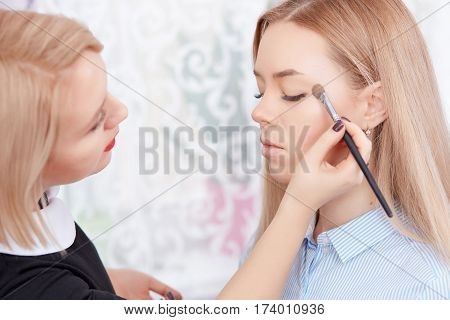 Side view of cosmetologist using long brush making makeup for attractive girl. Blonde woman client with plump lips and pure skin enjoying in beauty salon during procedure.