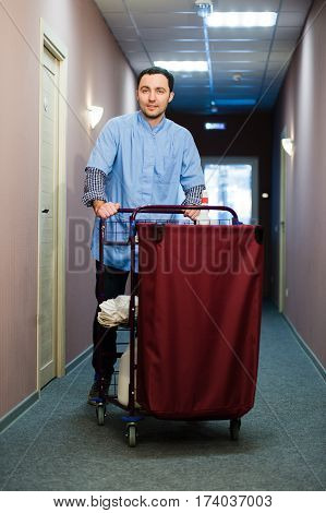 Young man pushing a housekeeping cart laden with clean towels, laundry and cleaning equipment in a hotel as he services the rooms.