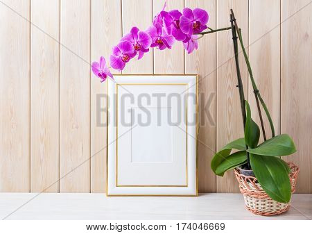 Gold decorated frame mockup with magenta-purple orchid in wicker basket near wooden wall. Empty frame mock up for presentation design. Rustic framing template for modern art.