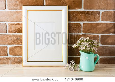 Gold decorated frame mockup with wild very soft pink flowers in mint pitcher near exposed brick wall. Empty frame mock up for presentation design. Template framing for modern art.