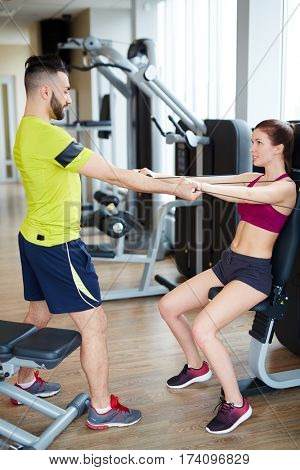 Working out in gym:  personal fitness instructor heping young woman do correct arm excercise on cable machine