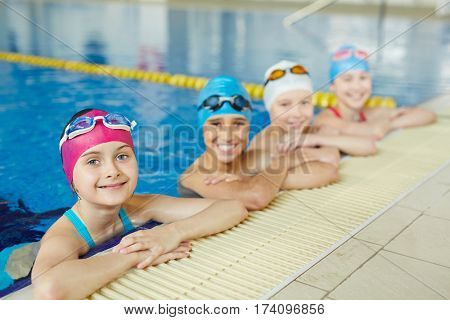 Group of cheerful school kids smiling looking to camera at border of swimming pool during practice
