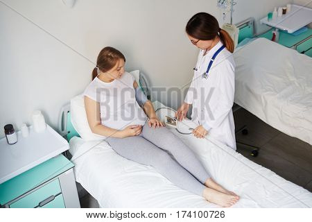 Female doctor measuring pressure of pregnant woman lying in hospital bed