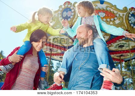 Carefree little girls playing with each other while sitting on shoulders of their smiling middle-aged parents, colorful merry-go-round observed on background