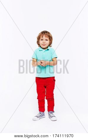 Full body shot of shy little boy dressed in bright colorful clothes standing smiling and twiddling hands against white background