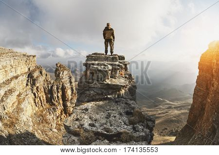 A young man stands on a rock surrounded by cliffs, located above the clouds in a great location, on a background of clouds, valleys, fields, mountains and sunset