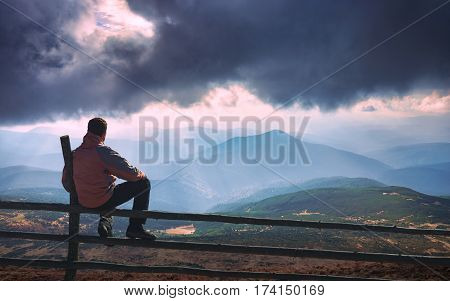 Man sitting on a wooden fence and enjoy rays of sunlight shining through the clouds to the mountain hills. Carpathian valley.