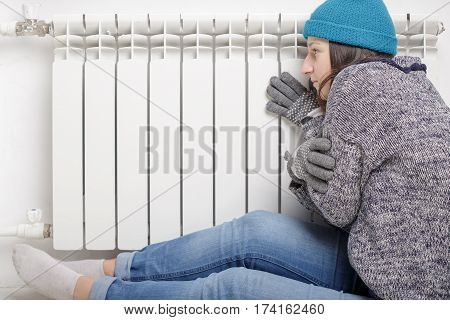 Young beautiful girl froze and warms hands near a radiator dressed in gloves a sweater and a hat