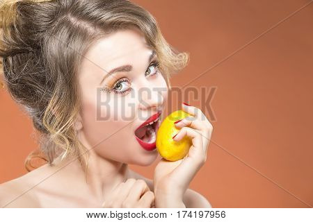 Sexy Fruit Series. Closeup Portrait of Happy Naked Caucasian Blond Girl Biting Yellow Lemon Fruit. Posing Against Orange Background. Horizontal Image