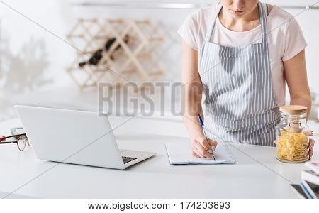 Noting the essentials. Focused dedicated pretty woman using her laptop finding a pasta recipe while making notes in her notebook