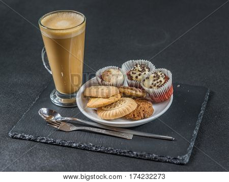 right side view on an aromatic coffee latte with three buttered muffins and petit fours on a white saucer with a spoon and forks the whole on a black stone plate sweet set