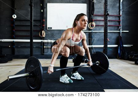 Intense workout in dark gym: bodybuilder woman ready to pick up heavy barbell with effort standing in squatting stance