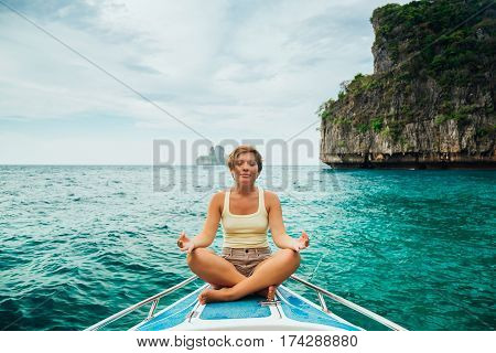 Beautiful girl with short hair in t-shirt and chert is sitting in a lotus position meditating on the deck of the yacht. Concept of calm and meditation.
