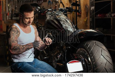 My iron friend. Positive delighted biker wearing casual clothes looking at instrument sitting in semi position next to big motorcycle