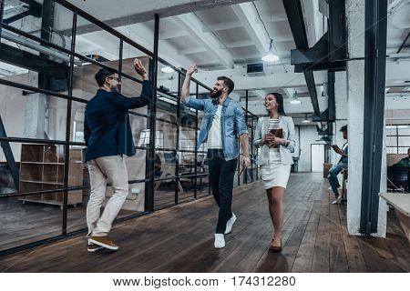 Give me five! Full length of two cheerful young business people giving high-five while their colleague looking at them and smiling