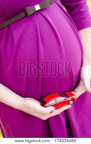 Pregnant in a purple dress holding a red toy car next to the tummy