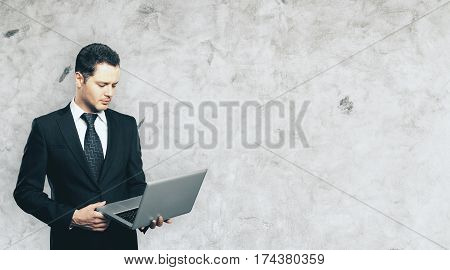 Handsome young businessman using laptop on concrete wall background with copy space. Technology concept