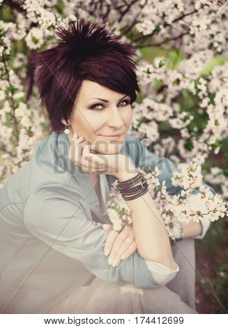 Young beautiful woman with short hair in blue jacket sitting near a bush blooming cherry. Spring. Romantic style