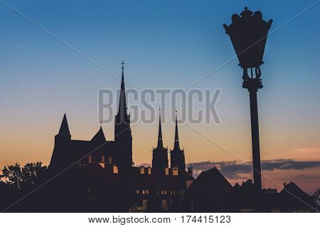 Wroclaw Cathedral island view from Oder river by sunset. Holy Cross church, St. John the Baptist cathedral and street lantern silhouettes by golden hour. Cathedral island in Wroclaw, Poland.