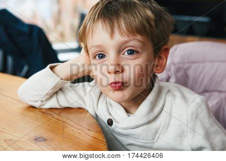 Portrait of cute adorable funny Caucasian little boy toddler in white shirt smiling making faces having fun sitting at table in restaurant indoors